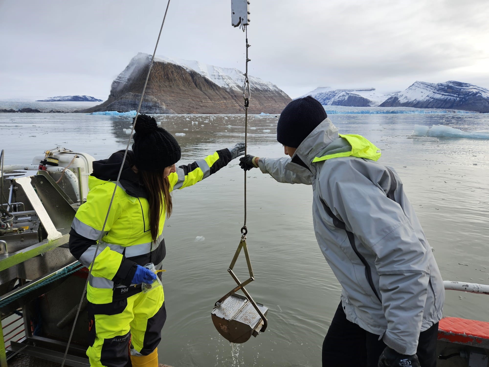 Two ICEBERG researchers lifting a sample collecting device from the ocean in an arctic landscape.
